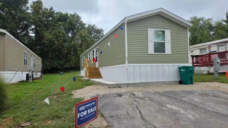 Single-wide mobile home with wooden entry stairs and a "For Sale" realty sign on grassy lot