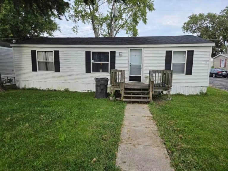 White mobile home with black shutters and wooden deck stairs leading to front door on grassy lawn
