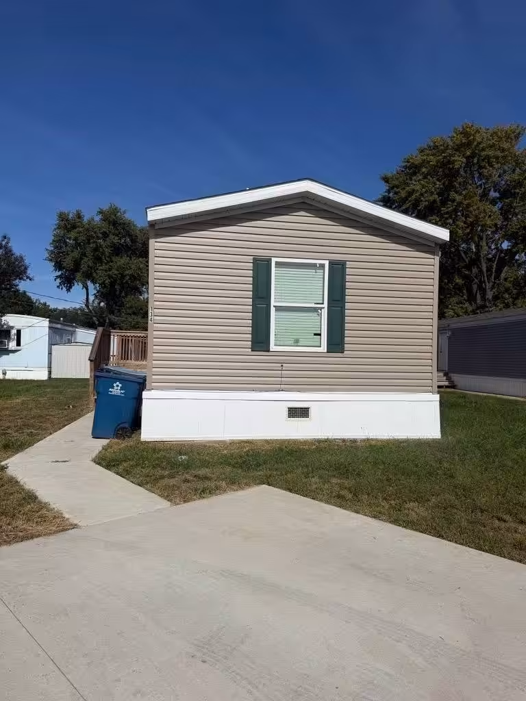 Tan mobile home with green shutters and blue recycling bin beside white skirting on grassy lot