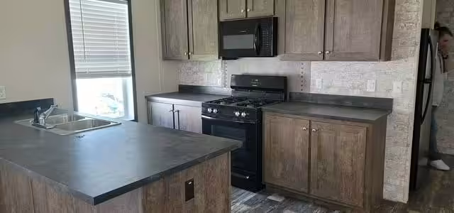 Kitchen with brown cabinets, black stove and microwave, dark island counter, and a window with blinds.
