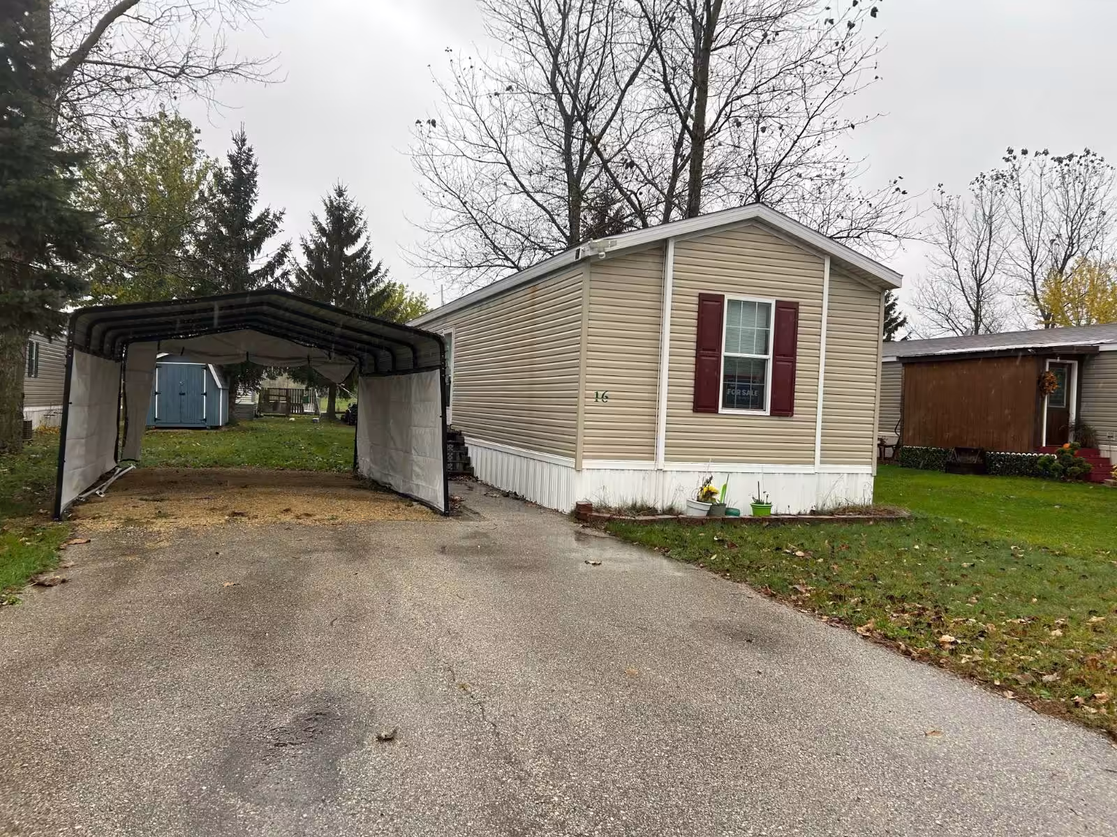Beige mobile home with maroon shutters, carport, and paved driveway, marked 16.